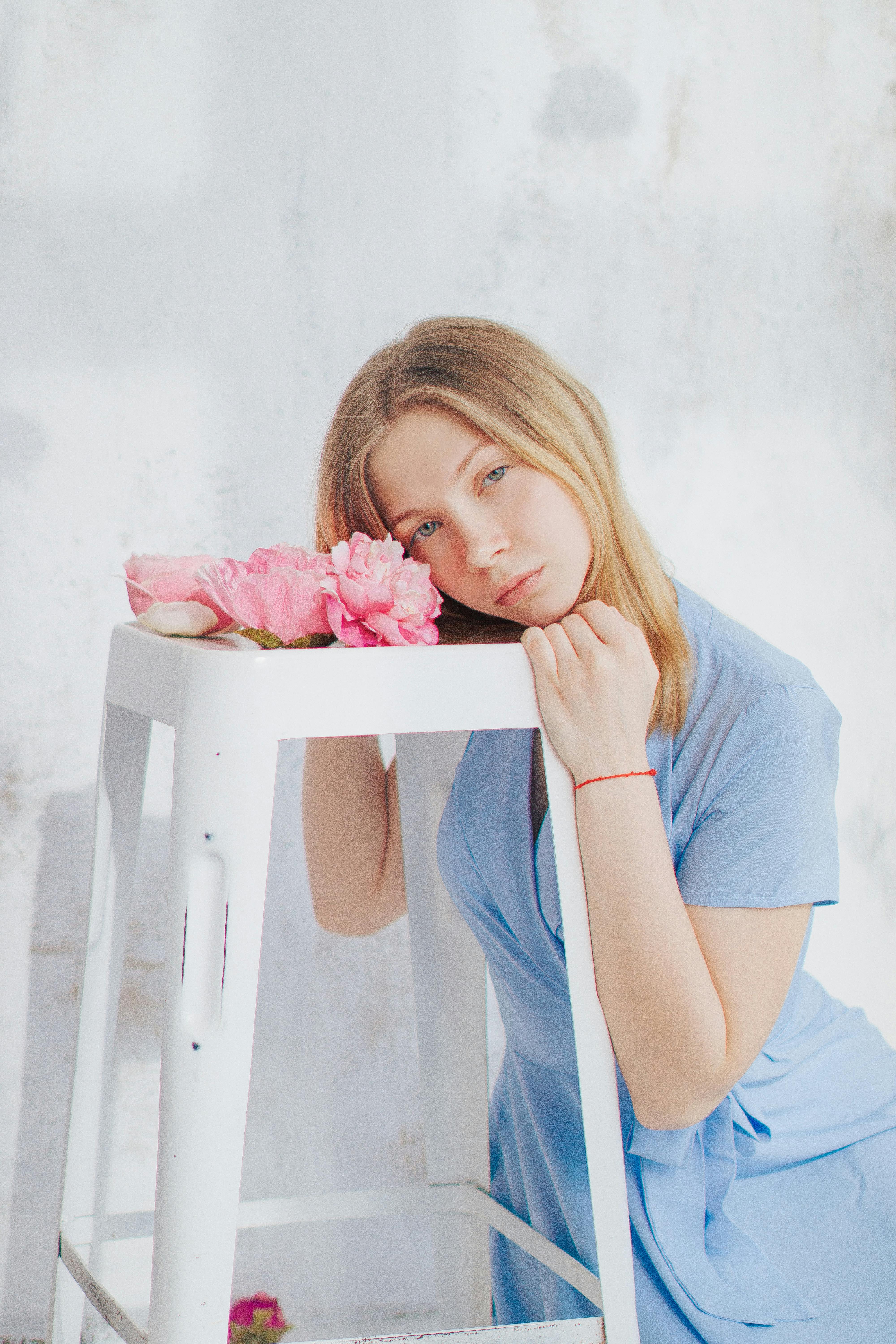 Young woman in a blue dress leaning on a white stool with pink flowers indoors.