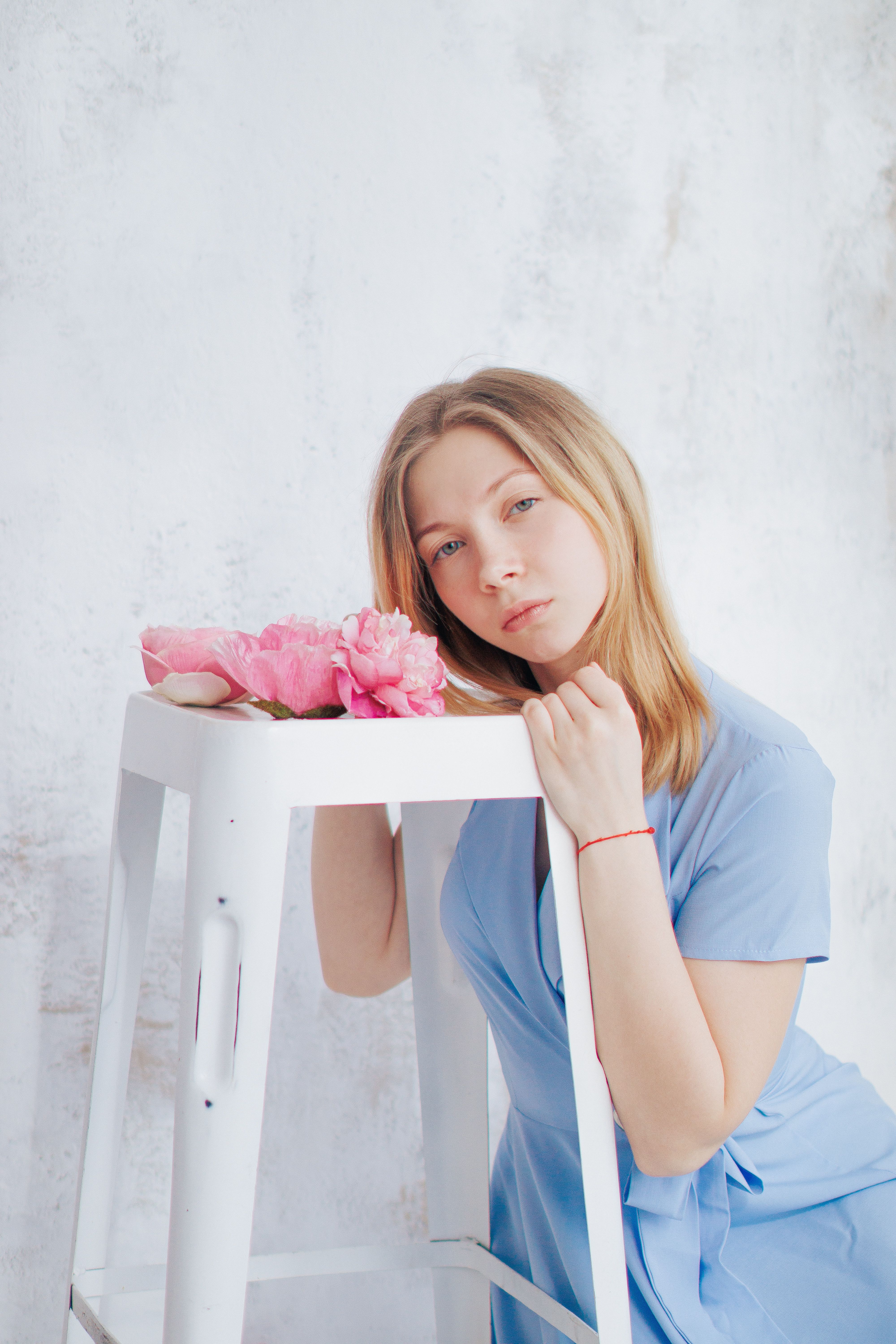 Young woman in a blue dress poses with pink flowers on a stool indoors.