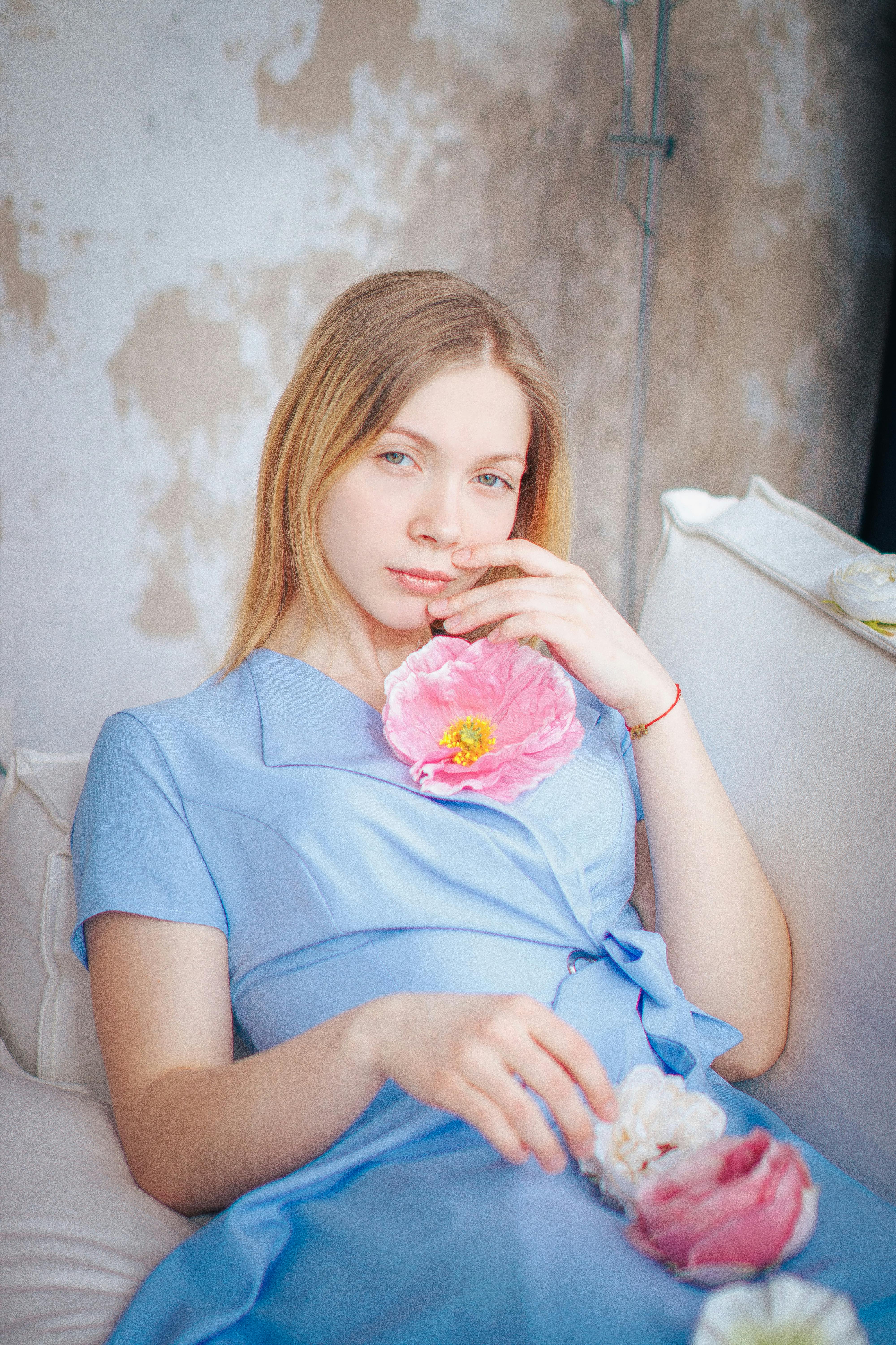 Young woman in blue dress relaxing on couch with pink flowers, exuding elegance and fashion.