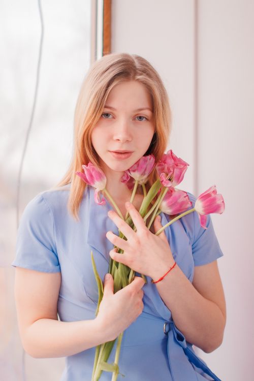 Free Blonde woman posing with pink tulips indoors, dressed in a blue outfit, in soft daylight. Stock Photo