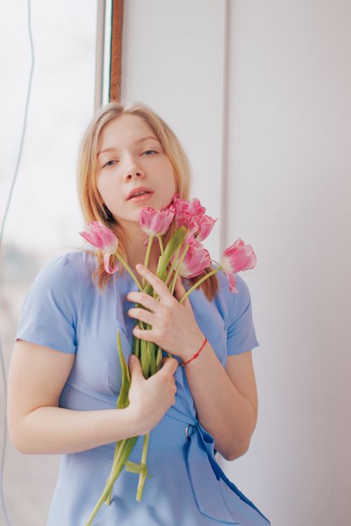 Free Young woman in a blue dress holding pink tulips by a window, conveying a soft and delicate mood. Stock Photo