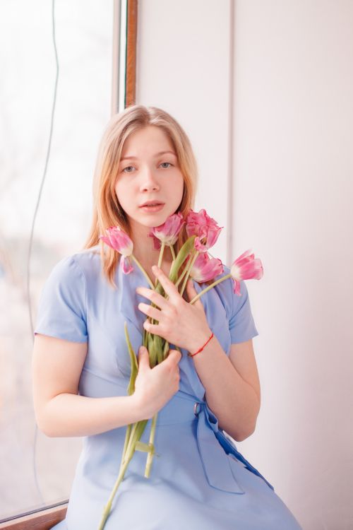 Free A serene woman in a blue dress holding pink tulips by a window, exuding tranquility. Stock Photo
