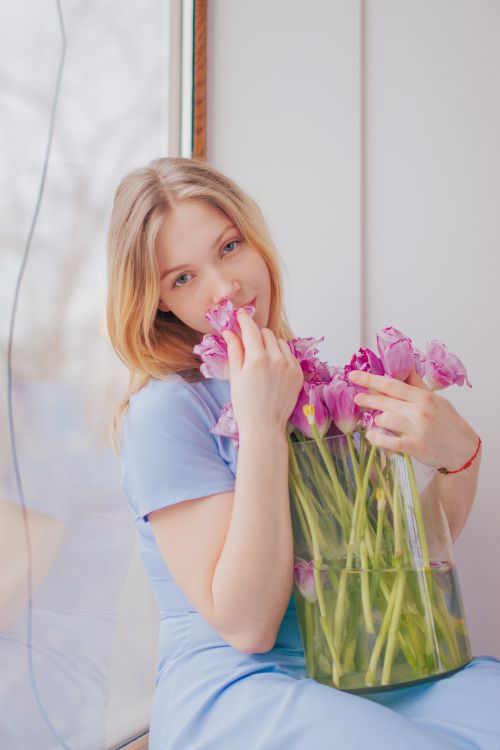 Free A young blonde woman in a blue dress holds a vase of pink tulips by a window, enjoying the view. Stock Photo