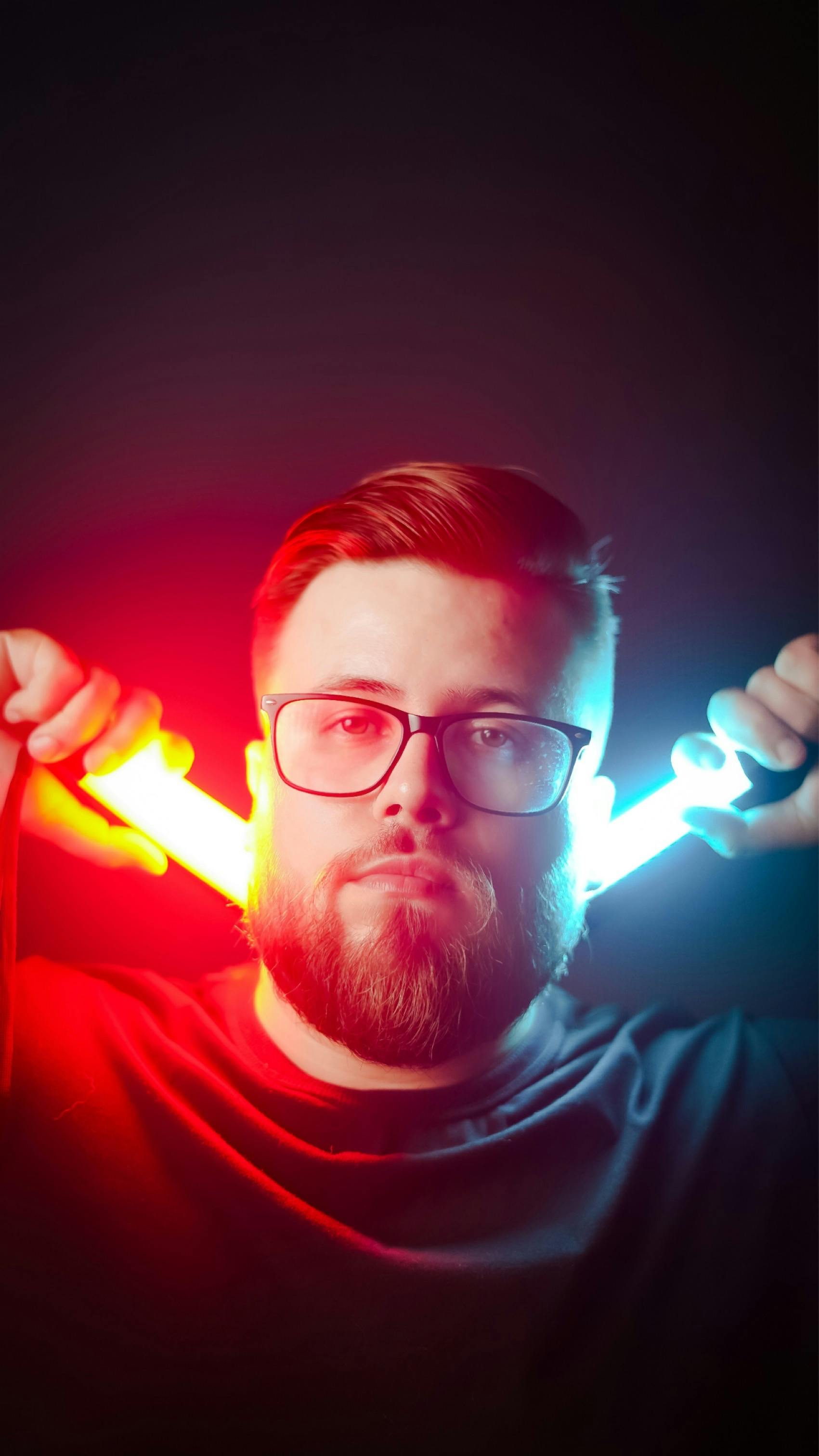 A bearded man with glasses holding neon lights in a dark room with vibrant colors.