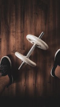 A close-up overhead view of a dumbbell on a wooden floor with feet in sneakers nearby.