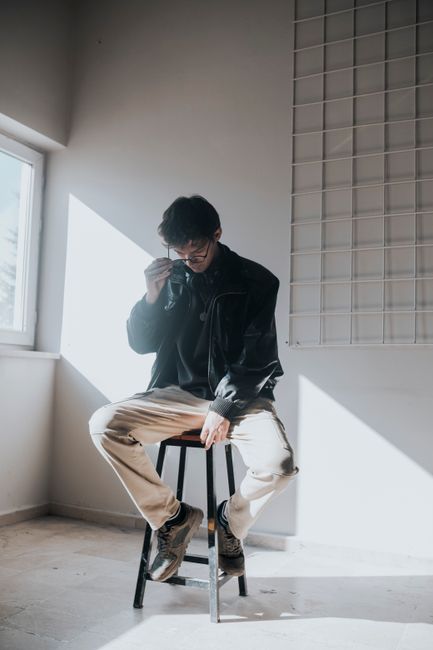 A man wearing eyeglasses and a jacket sits pensive on a stool in a sunlit minimalist room.