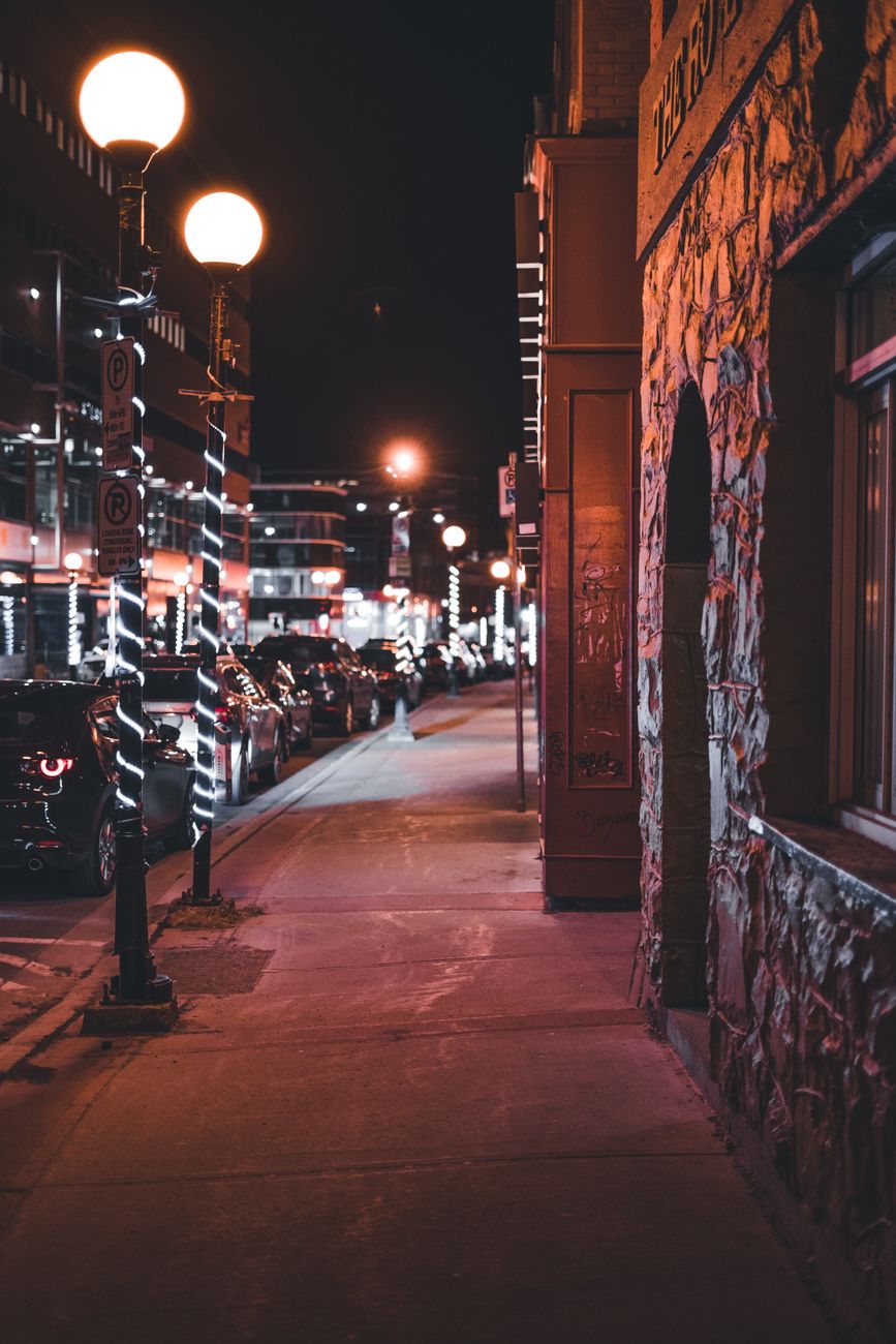 A lively nighttime urban street scene with glowing streetlights and parked cars on a city sidewalk.