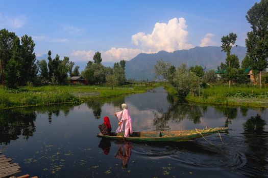 Two women rowing a narrow boat on a tranquil lake surrounded by lush greenery and mountains.