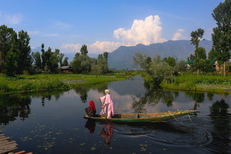 Two women rowing a narrow boat on a tranquil lake surrounded by lush greenery and mountains.