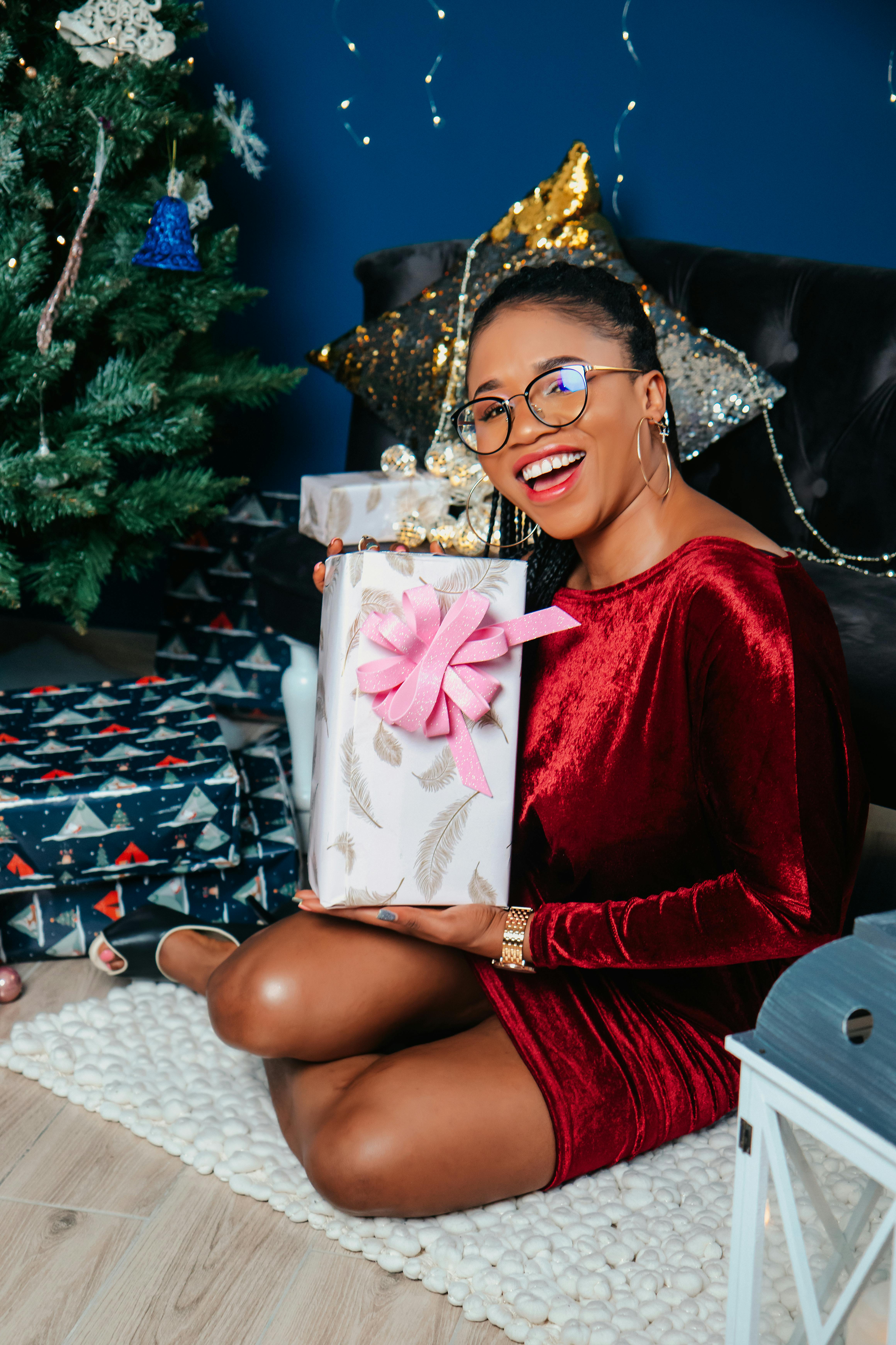 A joyful woman in a red dress holding a gift box by a Christmas tree.
