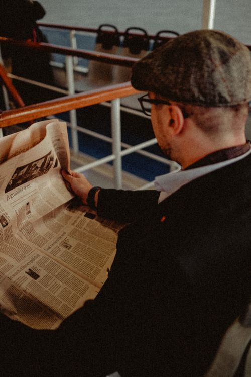 Free Back view of a man reading a newspaper indoors, wearing eyeglasses and a hat. Stock Photo