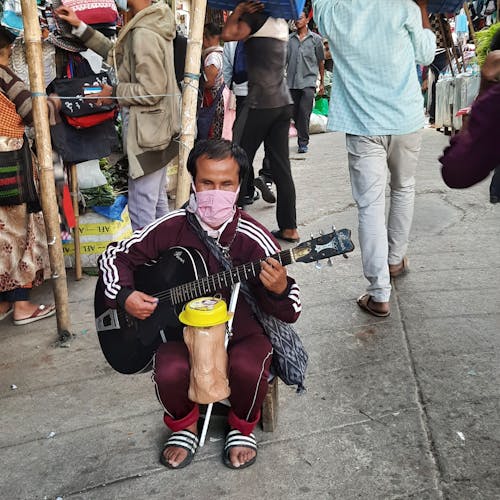 A physiscally blind handicappe man sitting on the roadside of a busy market playing a Guitar musical instrument in a way for colleting of donation from the general public.