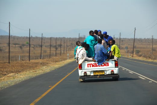 People riding on a Mazda pickup truck along a rural road under a clear sky.