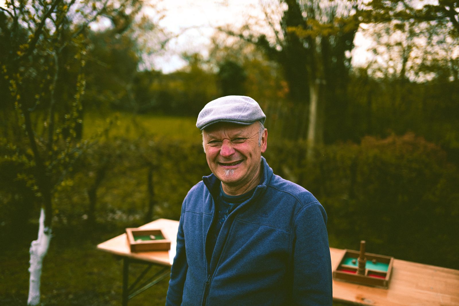 A smiling older man in a flat cap and navy fleece standing in a garden