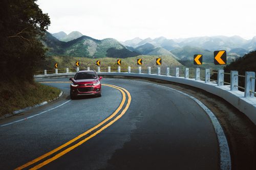 Free A red car drives along a winding mountain road with arrow signs and lush green scenery. Stock Photo
