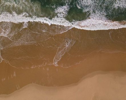Drone shot capturing the meeting of waves and a sandy beach shoreline from above.