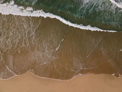 Free Stunning aerial view of ocean waves gently crashing onto a sandy beach shore. Stock Photo