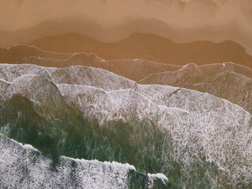 Free Aerial view capturing gentle ocean waves meeting the sandy beach in a serene and natural landscape. Stock Photo