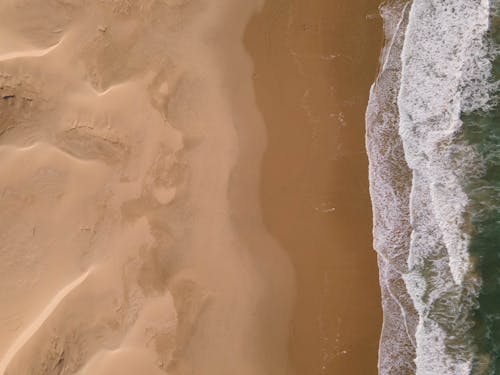 Drone image of green and blue ocean waves with foamy edges meeting a sandy yellow and brown beach.