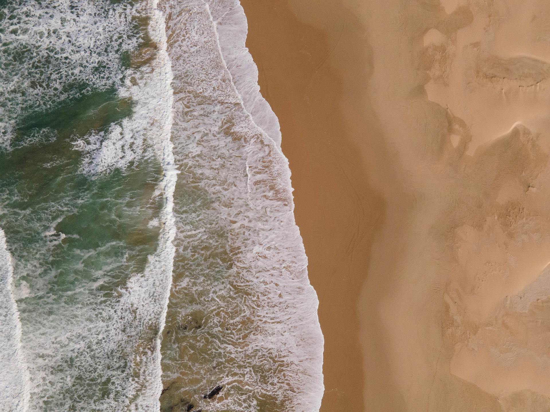 Top-down view of waves crashing onto a sandy shoreline, showcasing the texture and contrast.