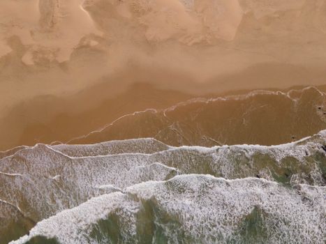 Drone's eye view of gentle waves lapping a golden sandy beach, creating a serene coastline scene.