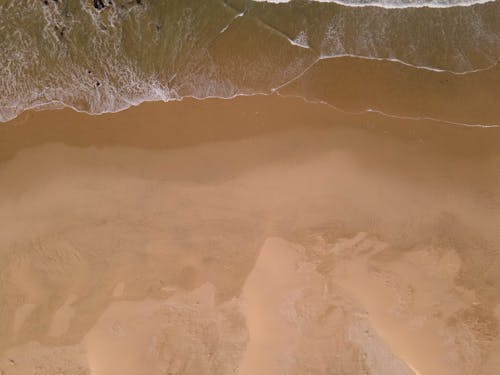 Drone image of green and blue ocean waves with foamy edges meeting a sandy yellow and brown beach.