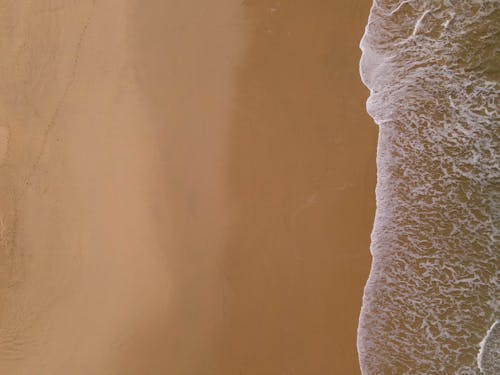 Drone image of green and blue ocean waves with foamy edges meeting a sandy yellow and brown beach.