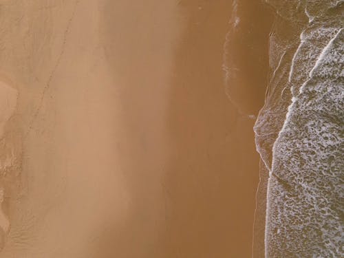 Drone image of green and blue ocean waves with foamy edges meeting a sandy yellow and brown beach.