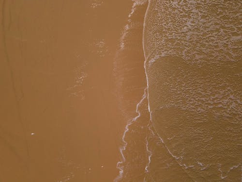 Drone image of green and blue ocean waves with foamy edges meeting a sandy yellow and brown beach.