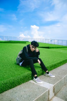 Casual portrait of a young man sitting outdoors on green grass under a clear sky.