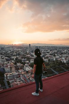 A man stands on a rooftop overlooking the Mexico City skyline during sunset, capturing the urban landscape.