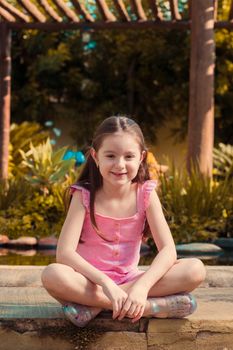 A young girl with long hair sits outdoors on a sunny day, smiling and wearing a pink outfit.