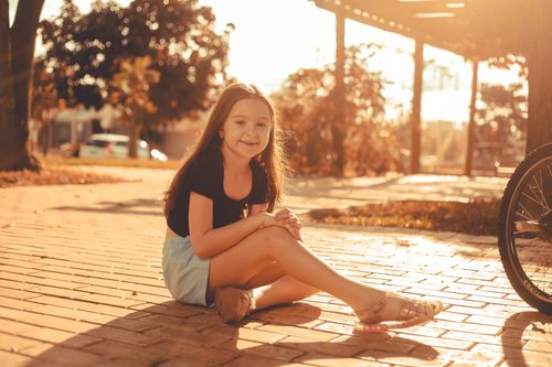 Free A young girl sits smiling on a sunlit sidewalk, exuding innocence and joy. Stock Photo