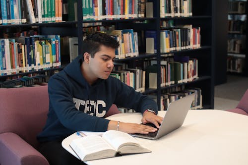 A Student Sitting in a Library and with a Laptop and Studying 