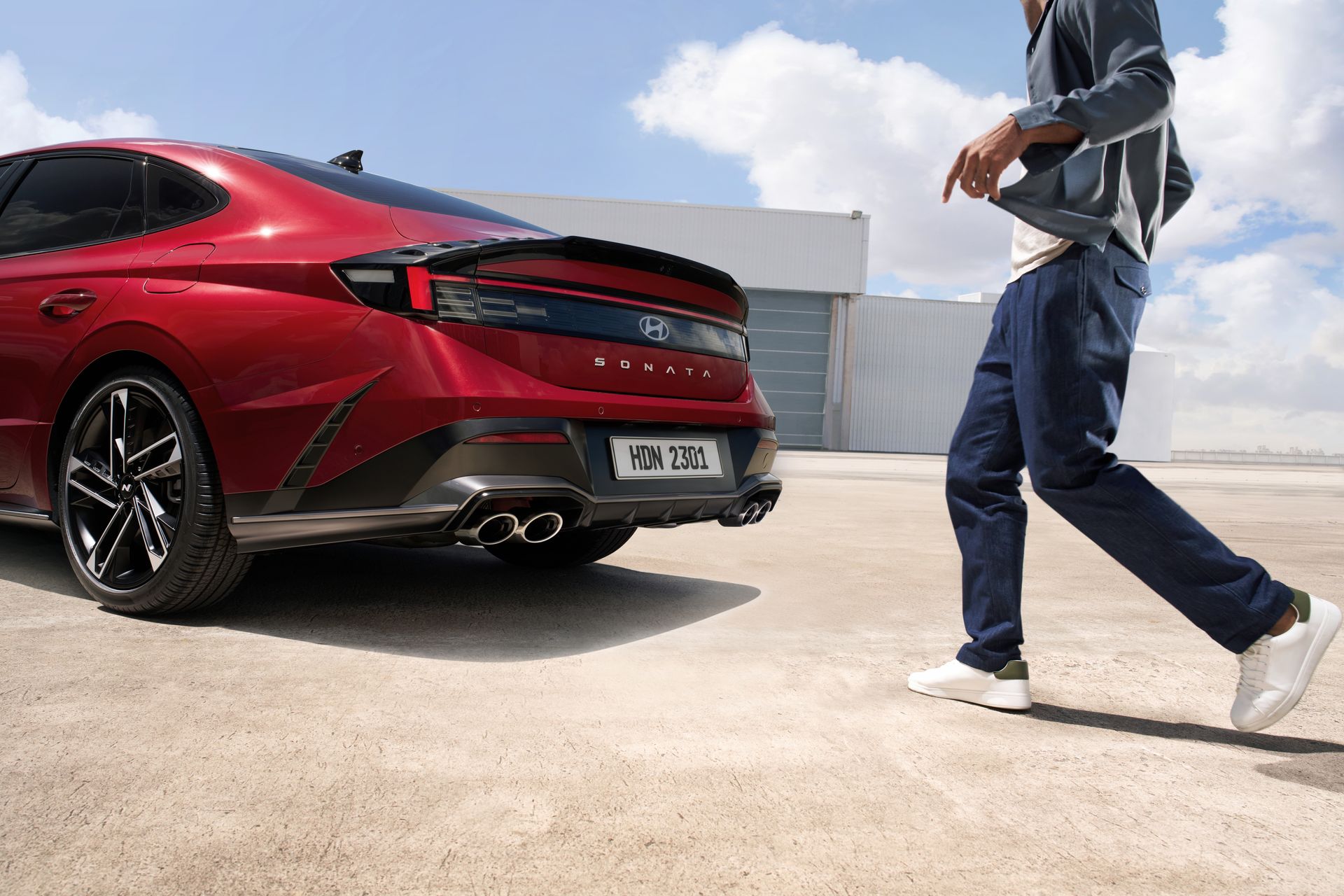 Dynamic shot of a red Hyundai Sonata N Line with a man walking nearby in a sunny parking lot.