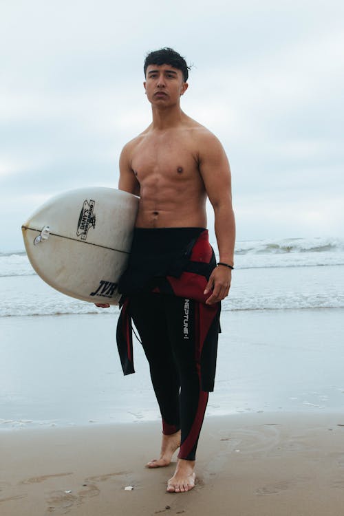 Young Man in a Wetsuit Holding a Surfboard on the Beach