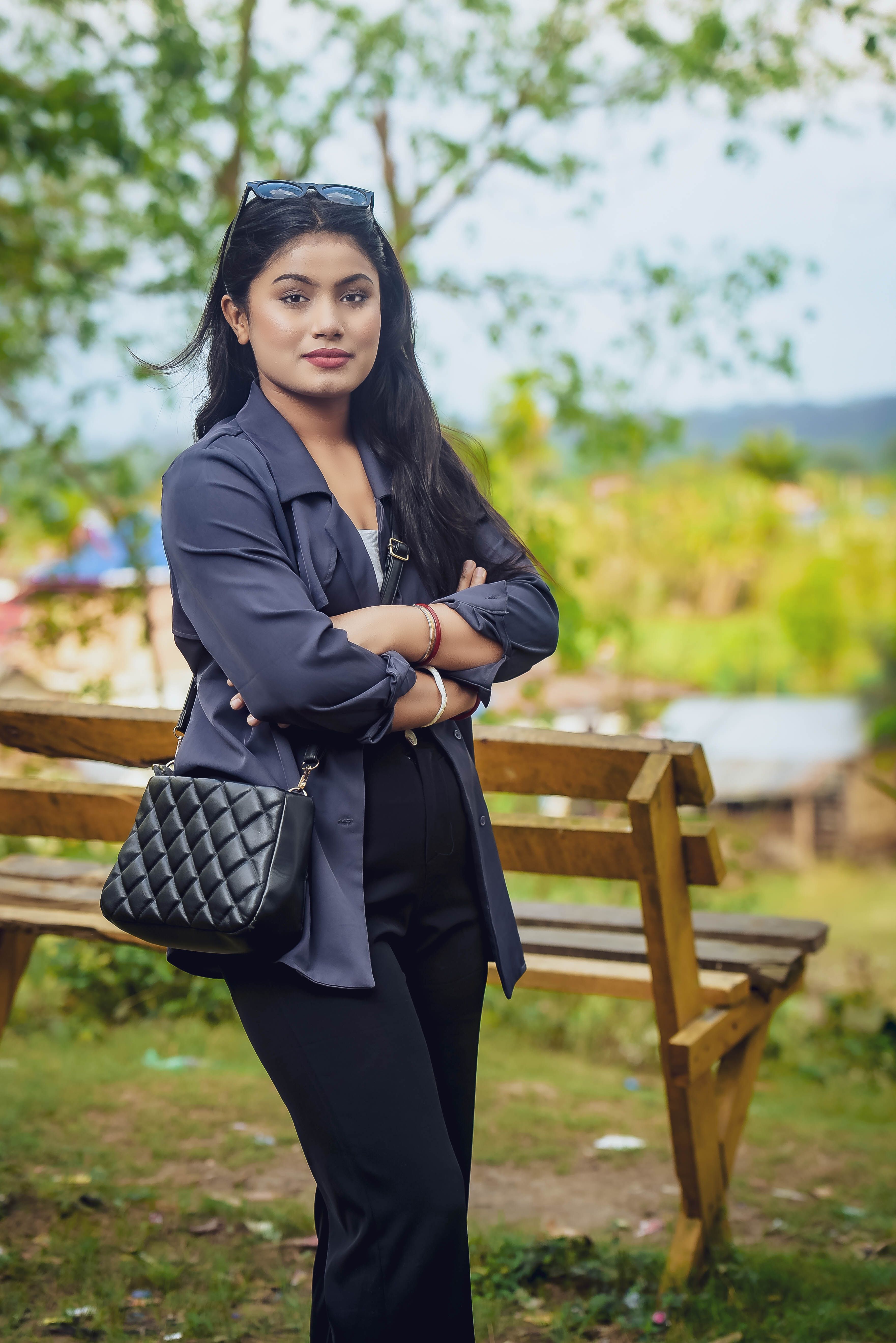 Elegant young woman in fashionable outfit posing by a bench in a scenic outdoor park.