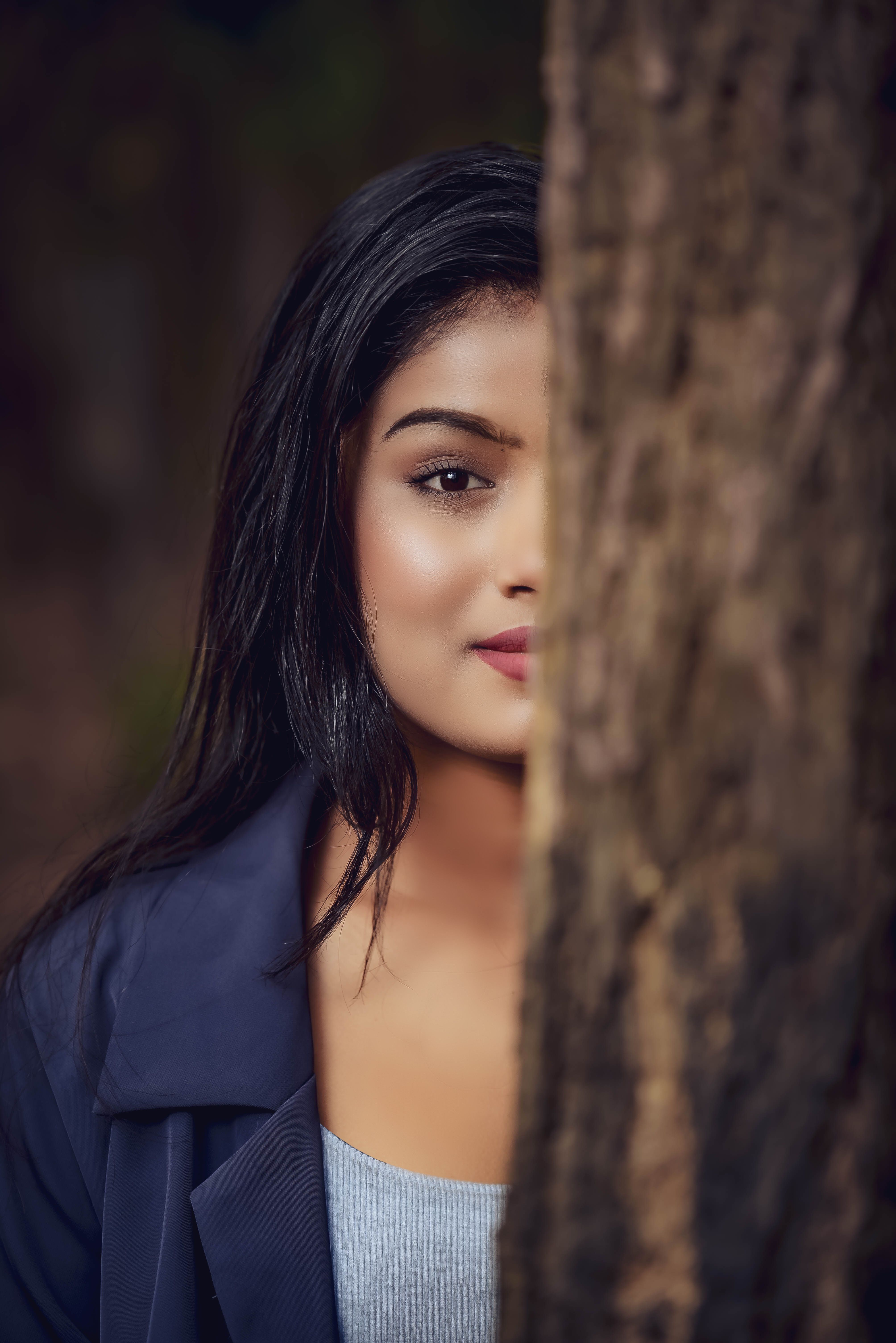 A young woman peeking behind a tree with selective focus in a casual outdoor setting.
