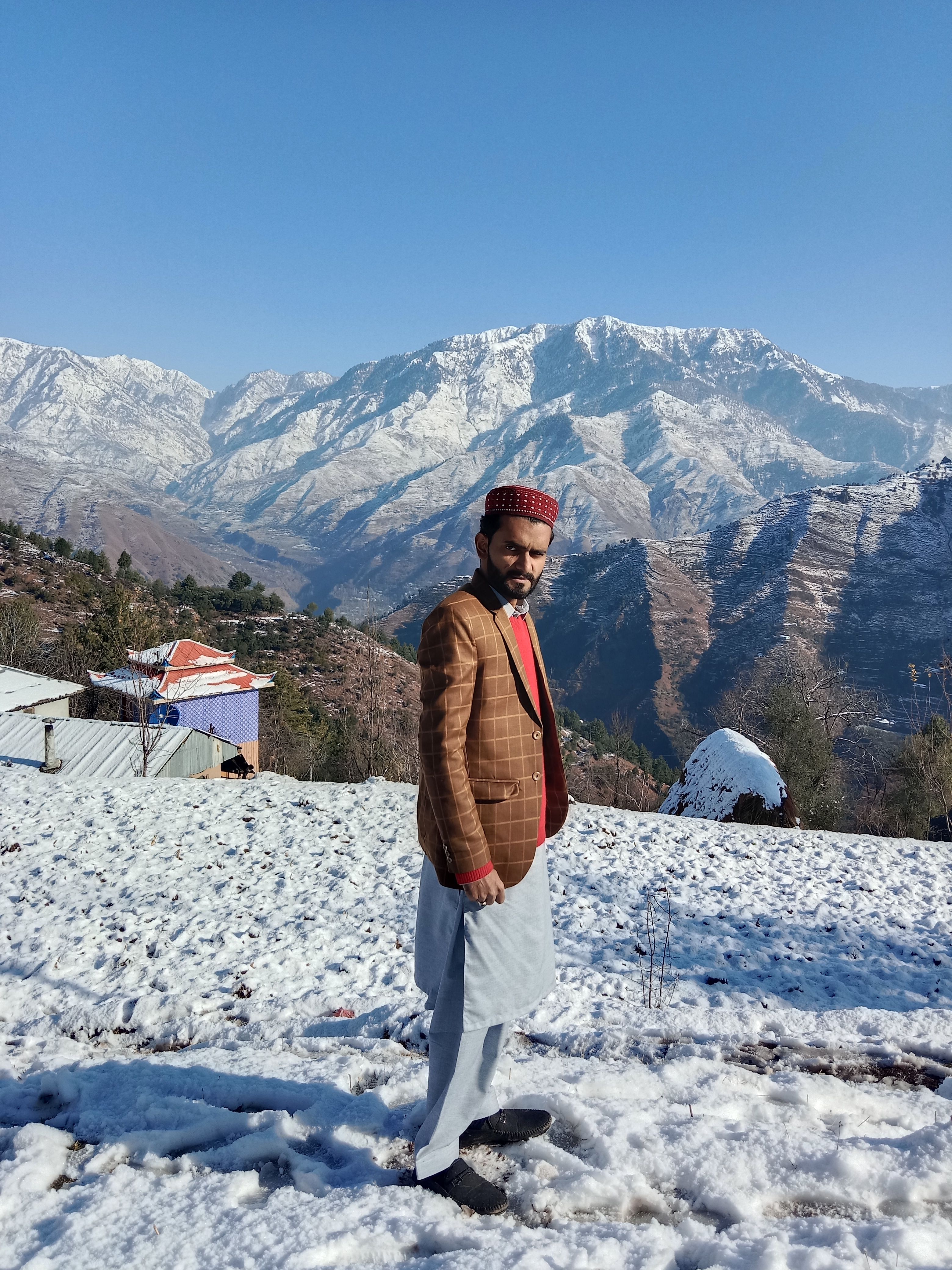 Man in a plaid jacket and traditional hat poses in snowy mountains, capturing winter fashion and scenery.