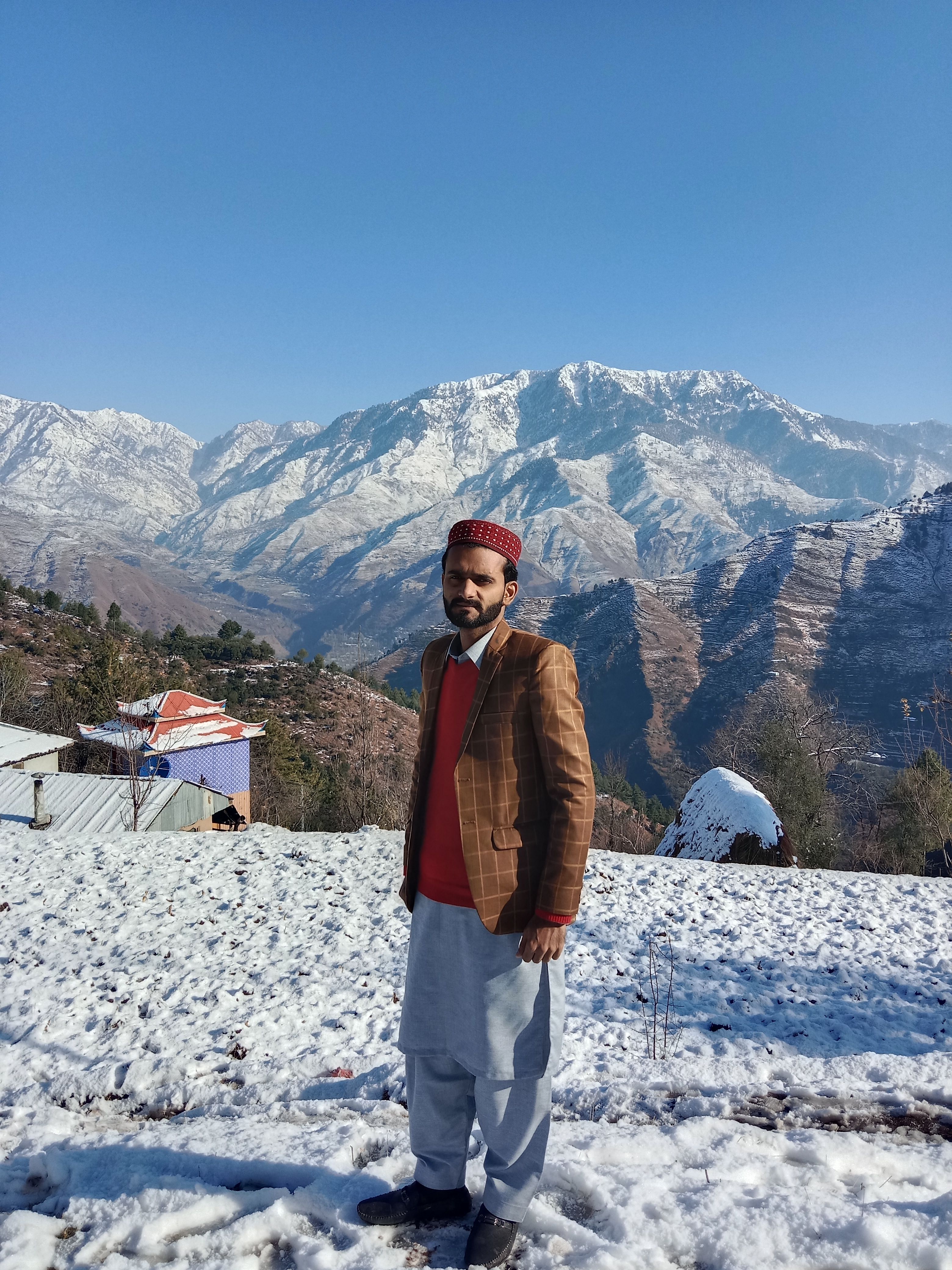 A man stands in traditional attire before sunlit snow-covered mountains.