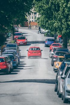 A sunlit urban street with parked cars and a red car driving, under clear sky.