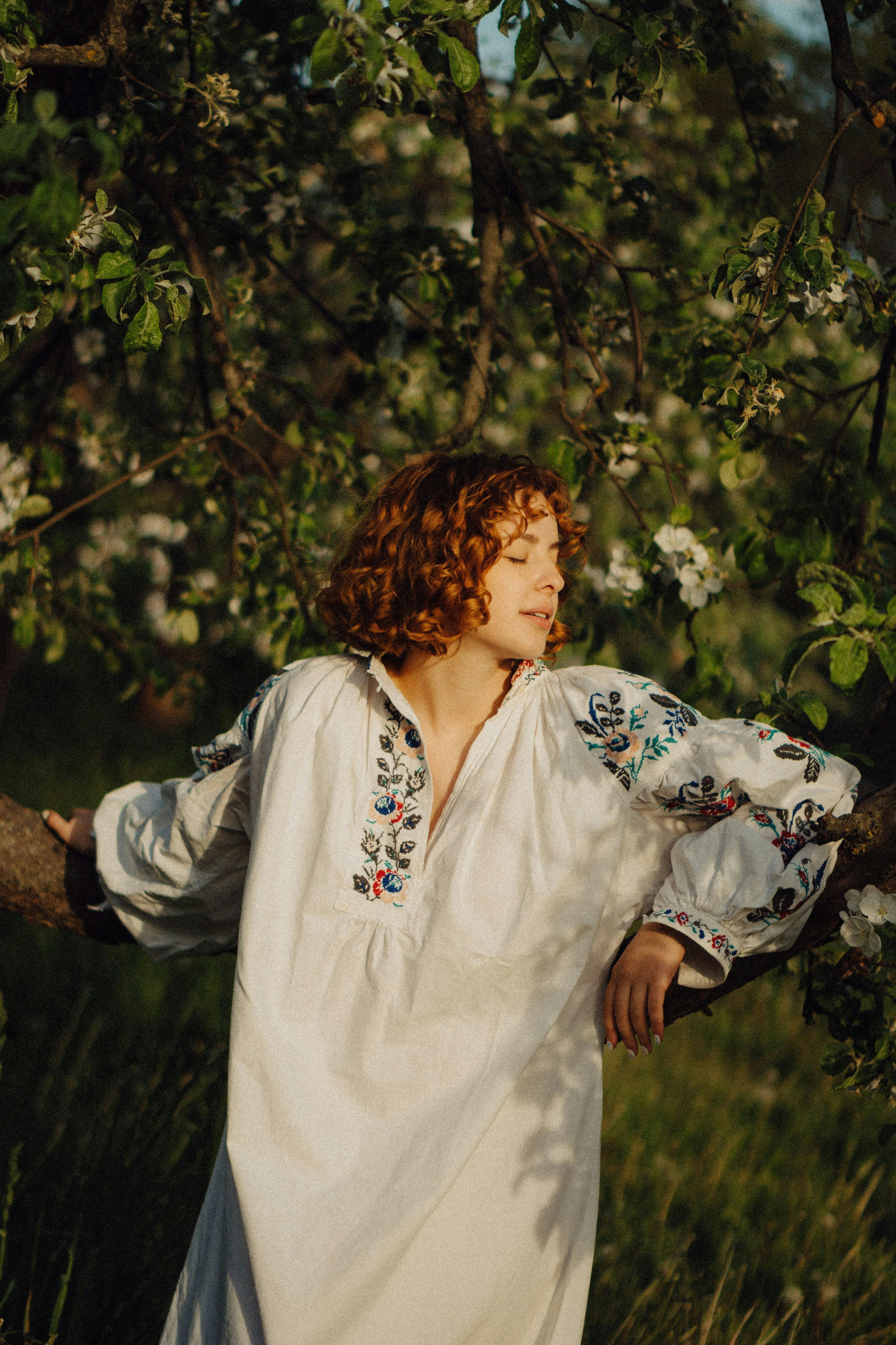 A young woman with curly hair enjoys nature in a sunlit orchard, wearing an embroidered dress.