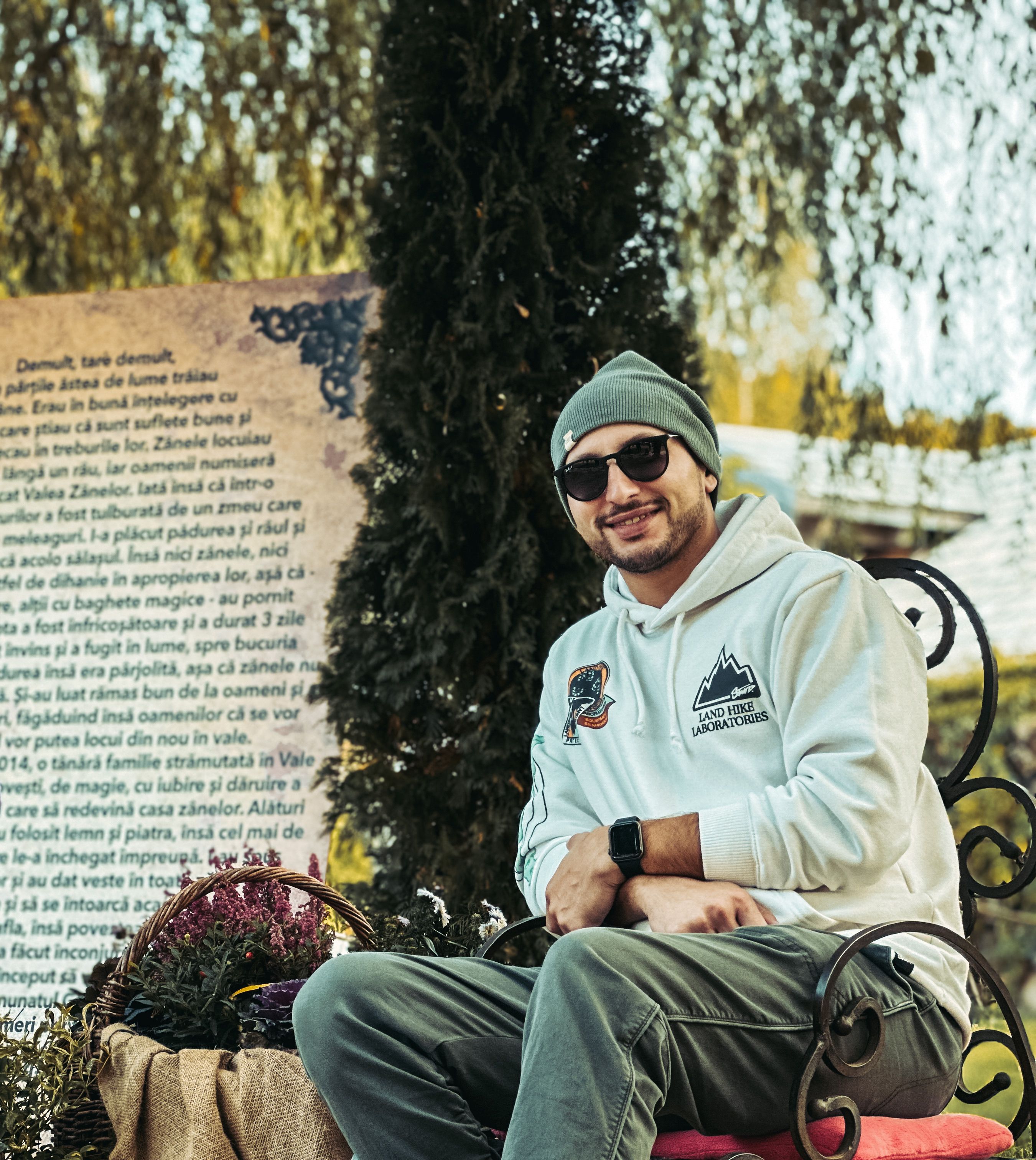 Smiling man in casual attire, wearing a hoodie and hat, sitting outdoors with a backdrop of trees.