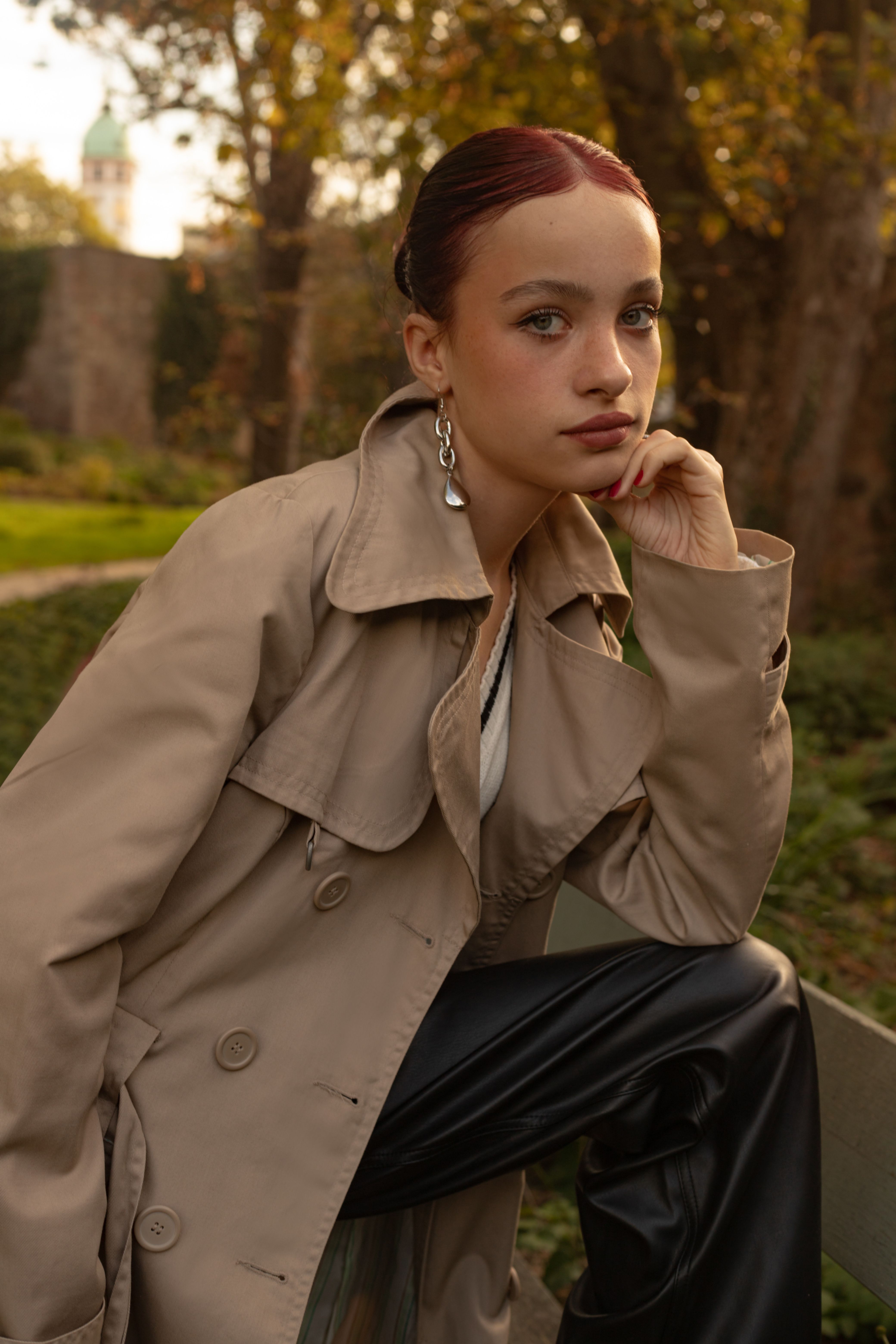 Fashionable woman posing in a trench coat outdoors, showcasing modern style under autumn light.