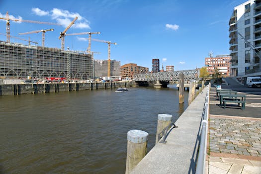 A vibrant cityscape with construction cranes, a river, and modern buildings under a clear blue sky.