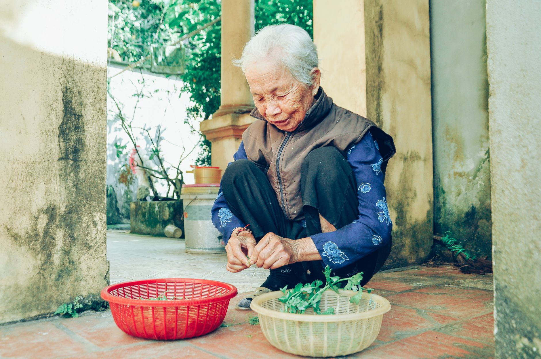 A senior woman sits outdoors, processing vegetables in baskets on a sunny day.