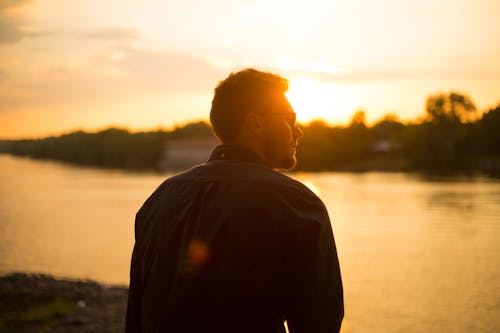 Free Man Sitting Near Large Body of Water Under Clear Sky during Sunset Stock Photo