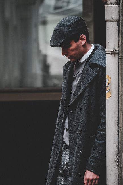 A fashionable man wearing a grey overcoat and flat cap leans against a wall in an urban setting.