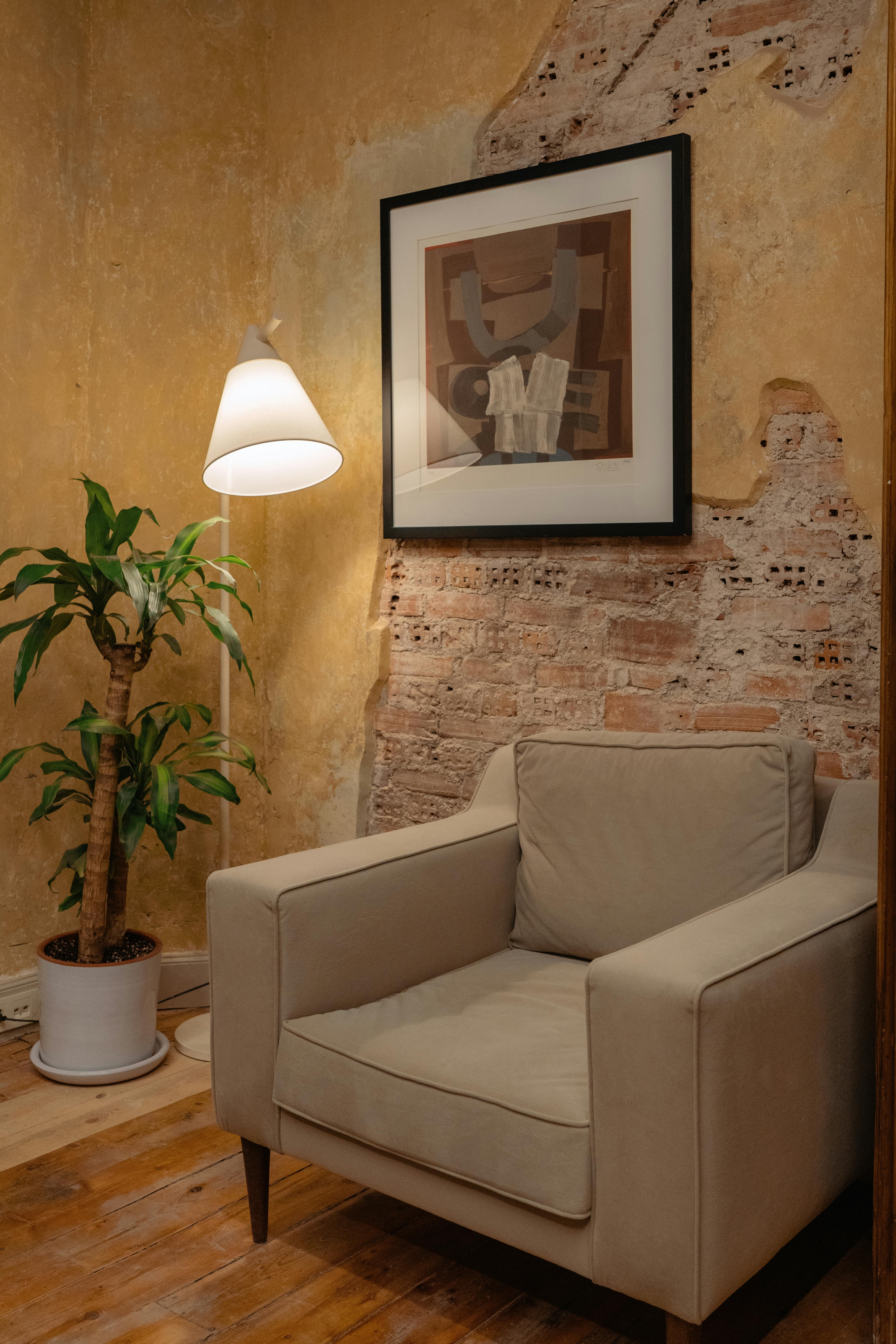 Free A serene corner with an armchair, potted plant, and artwork on a textured wall, illuminated by a chic lamp. Stock Photo