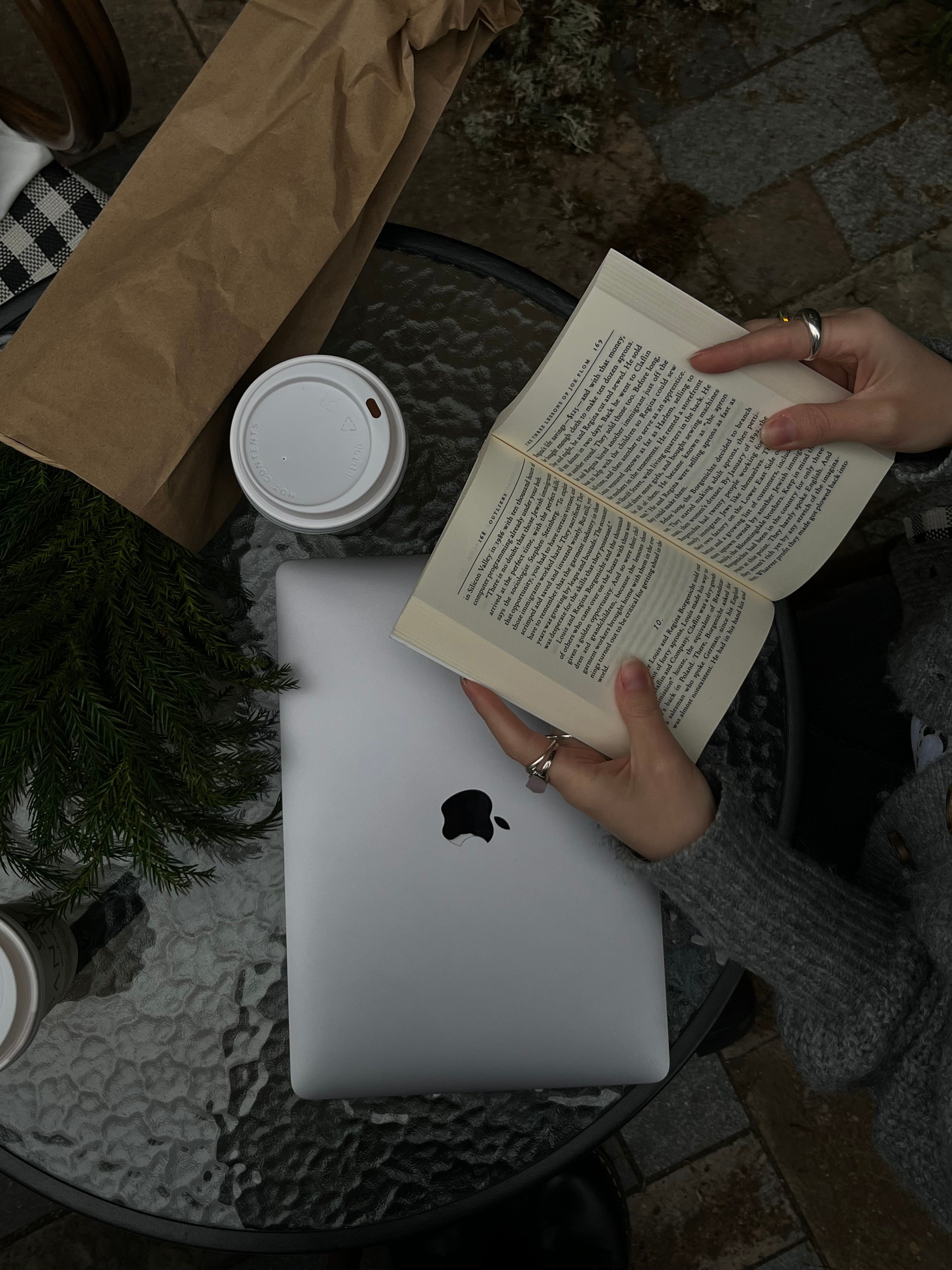 A person reading a book next to a laptop and coffee cup on a glass table, outdoors.